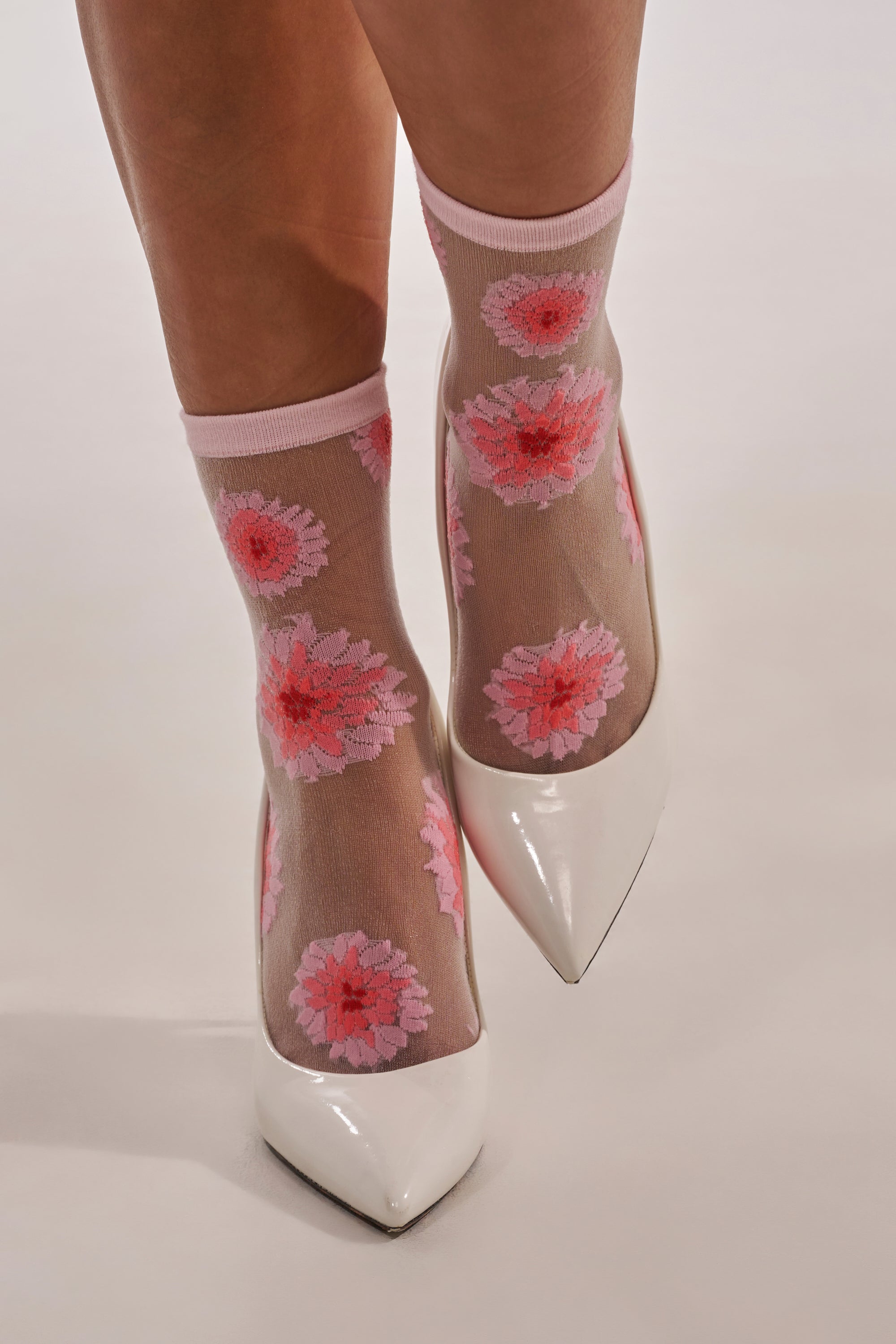 A person models WHERES MY HEEL SOCKS—cute mesh socks with pink and red floral patterns—paired with white pointed-toe heels on a light surface. 1