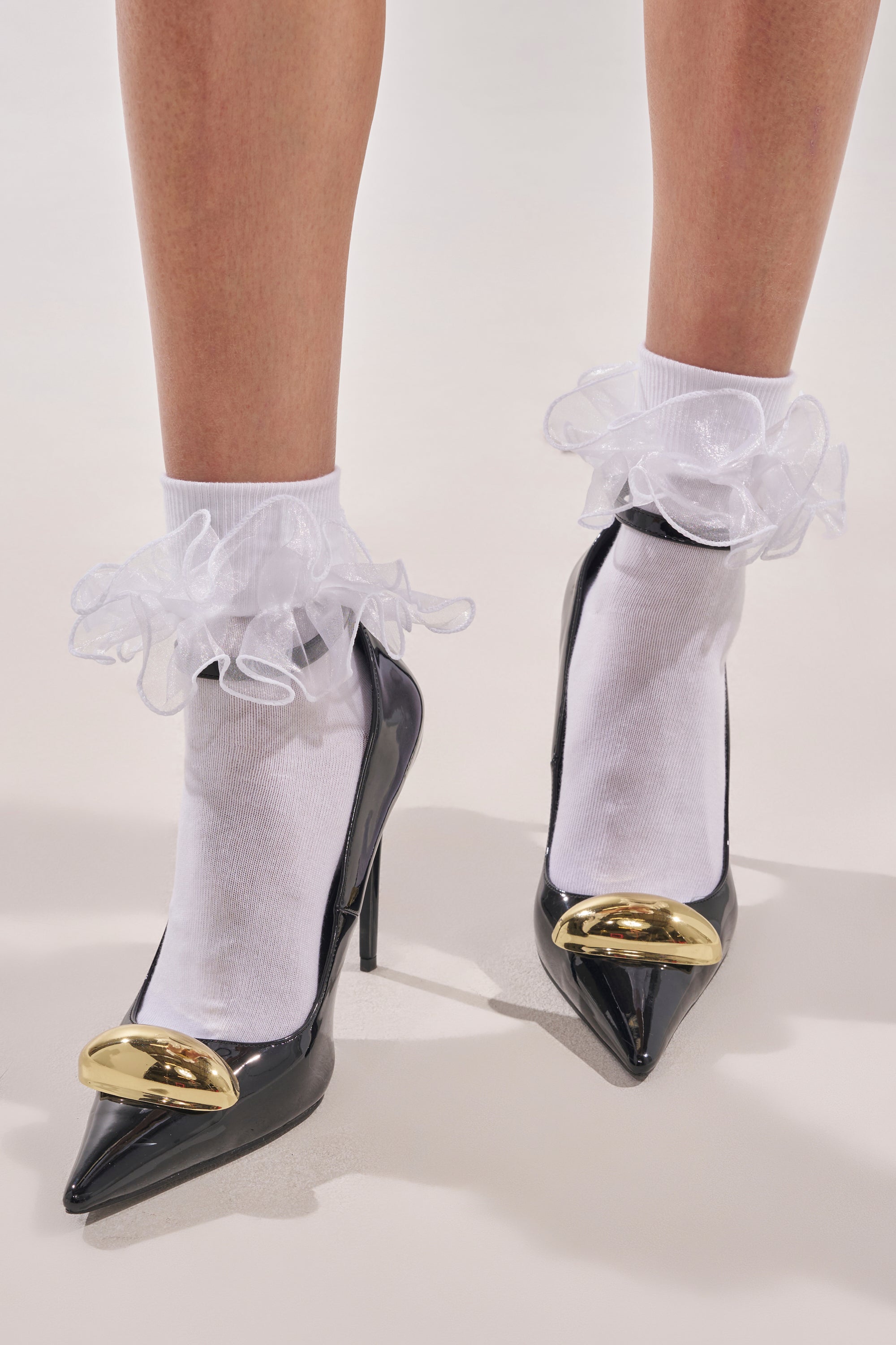 Model wearing RUFFLE ME UP SOCKS with frilly, ruffled lace trim, paired with black high-heeled shoes featuring gold pointed toe caps. Set against a plain light-colored background. 0