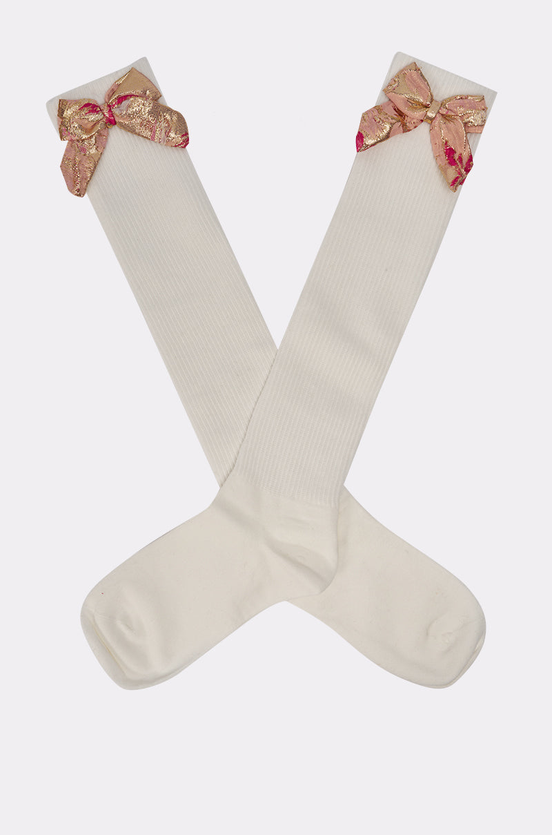 DOLLED UP BOW SOCKS: Ivory knee-high socks featuring large, shimmery gold and pink brocade bows near the top, shown crossed on a plain light background. 3