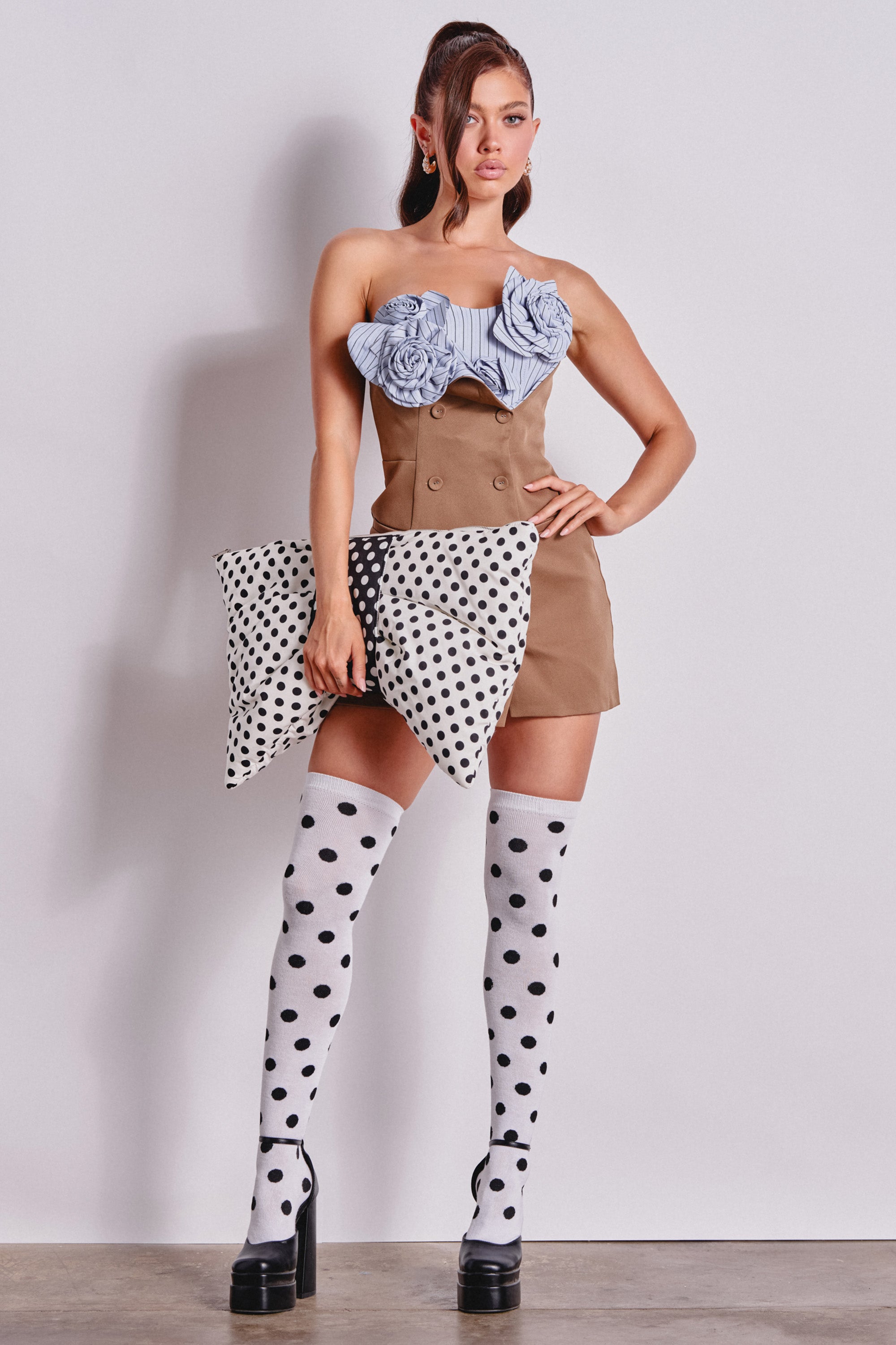 A woman poses confidently against a plain background in a brown mini dress and black platform heels, holding a polka dot pillow. She stands out wearing the playful CALL ME DOTTIE KNEE HIGH SOCKS with bold polka dot design. 0