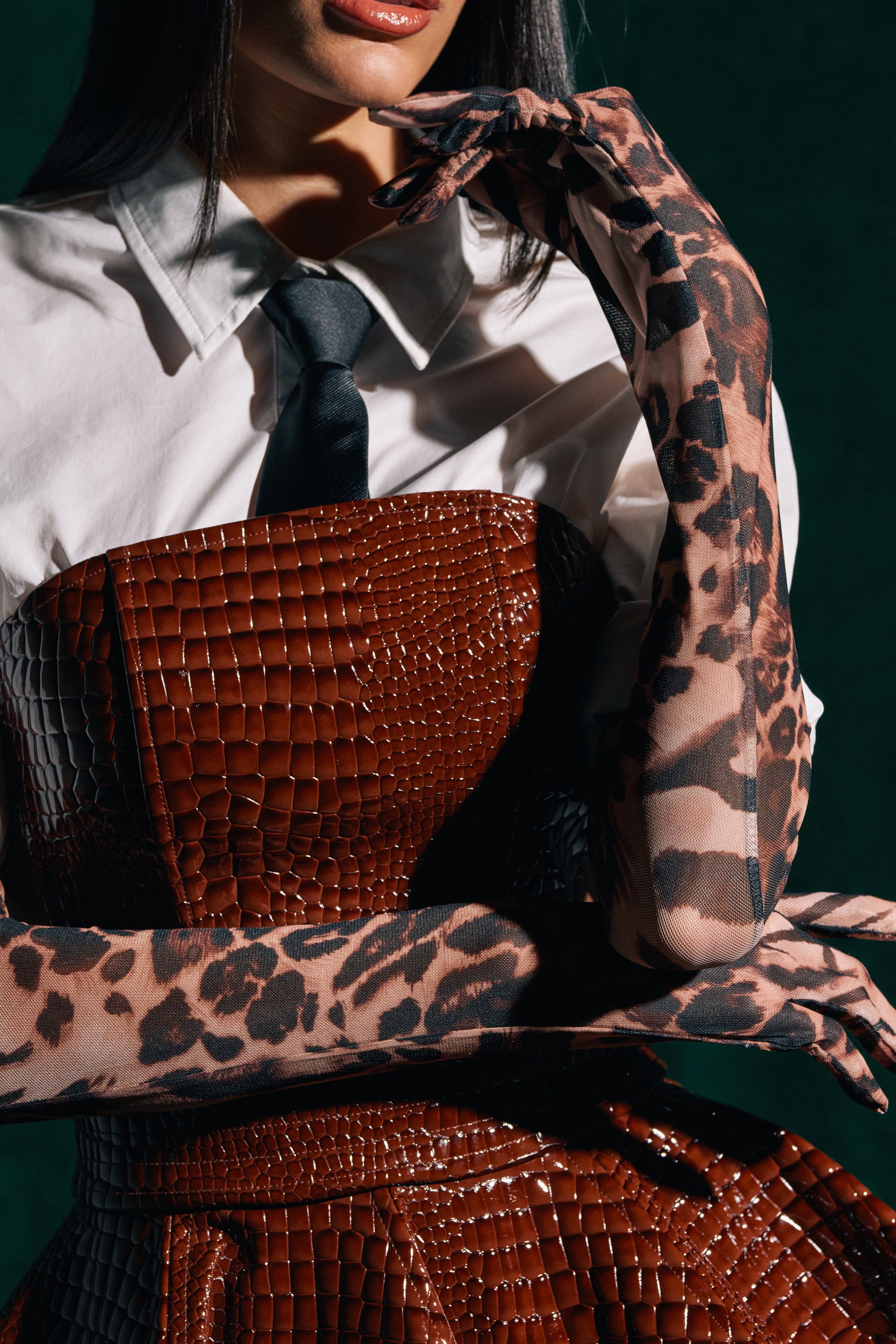 A woman poses with one hand near her face and the other on her waist, wearing a white collared shirt, black tie, brown faux crocodile corset dress, and REALLY WITH IT CHEETAH GLOVES.