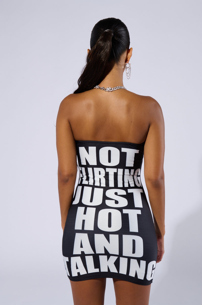A woman with a ponytail is dressed in the JUST HOT GRAPHIC MINI DRESS, a strapless mini dress with a bodycon fit, featuring bold white text that reads, "NOT FLIRTING JUST HOT AND TALKING." She complements her look with hoop earrings and a chain necklace. The plain, light-colored background accentuates her stylish attire beautifully.