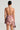 A woman is seen from behind wearing the SO Y2K MESH PRINTED MINI DRESS, featuring a backless halter neck design with an abstract brown and pink pattern, evocative of early 2000s fashion. The dress boasts ruffled layers at the hem. She accessorizes with a small purse, her hair styled neatly in a bun, and large earrings.