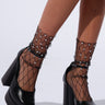 A person showcasing the PAIR ME DECORATIVE SOCKS—black fishnet socks adorned with small rhinestone embellishments—paired with black platform heels. The plain, lightly colored background highlights the decorative socks and footwear beautifully.