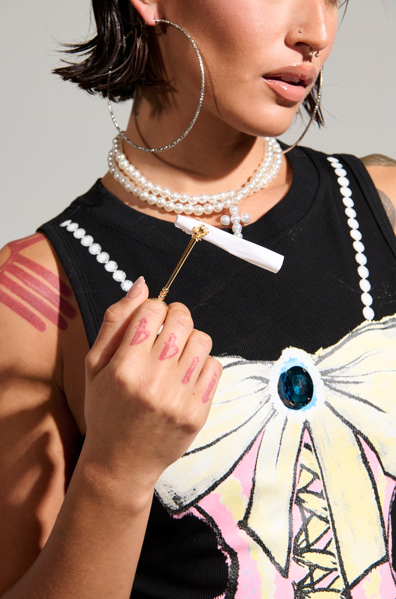 A person with short hair is wearing a black top featuring a decorative bow and an eye-catching large blue gem. They accessorize with a white stick topped with flowers, flaunt substantial hoop earrings, and showcase layered pearl necklaces adorned with a rose pendant. Additionally, they wear the HOLD IT RING IN GOLD on their hand which also displays lipstick marks alongside their arm. 2