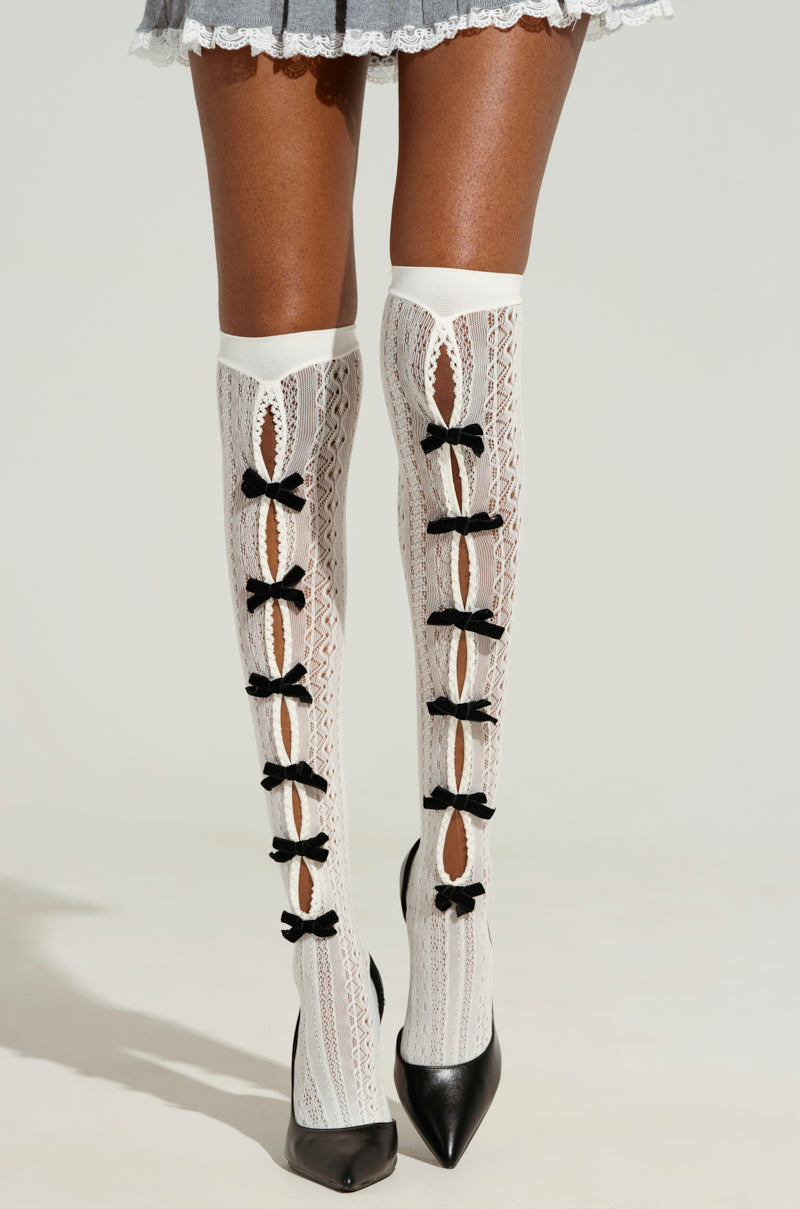 A person is wearing the HERE FOR THE VIBES SOCKS, which are white with a lace pattern and adorned with vertical velvet bow appliques down the back. They pair these socks with black pointed high heels and a short skirt featuring white lace trim.