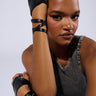 A person poses with their elbow resting on a surface and hand on their forehead. They are wearing a sleeveless top and the WRAPPED AROUND PU FINGERLESS GLOVES, which are black faux leather gloves that extend and wrap around their arms. These gloves add a touch of edgy biker accessory style to the scene. The background is plain and light-colored.