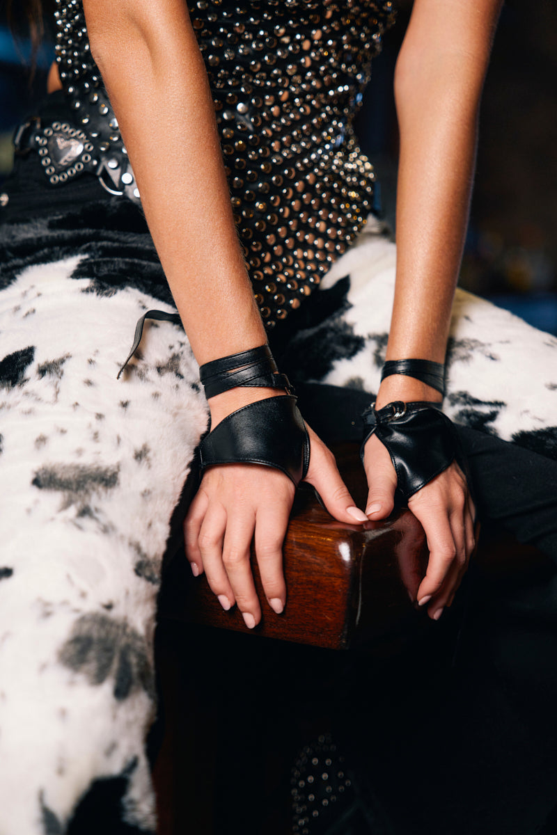 A person sits on a wooden surface wearing black-and-white cow print pants, a studded top, and WRAPPED AROUND PU FINGERLESS GLOVES with leather wrist cuffs, their hands resting in front.