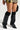 The individual sporting tall black leather boots with stiletto heels and LAYERED UP LEGWARMERS exudes a cozy vibe while wearing a snakeskin-patterned mini skirt. In their left hand, they hold a bag against a neutral background.