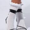 Close-up of feet wearing GIRLY RUFFLE SOCKS, white and textured with frilly edges, paired with black strappy high-heeled sandals that wrap around the ankles against a solid white background.