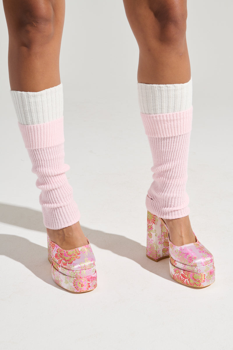 A person is standing and wearing the "WITH THE TREND LEG WARMERS," which feature a white and light pink knit fabrication, paired with high-heeled platform shoes adorned with a floral pattern. The background is a plain white surface.