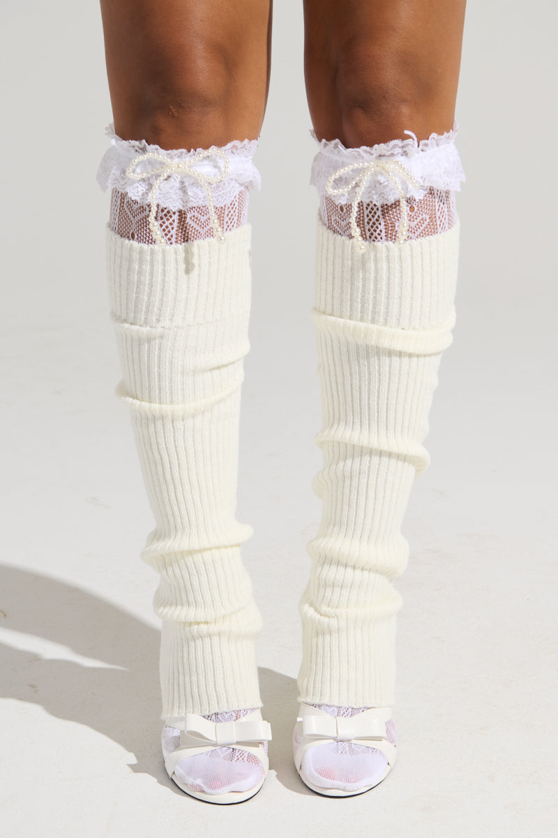 A person is shown from the knees down wearing COCO STOCKING LEG WARMERS in a cream-colored knit design, accented with lace trim at the top. The leg warmers are paired with white open-toe high heels featuring front bows. Small pearls add an extra layer of elegance to their Balletcore-inspired look. The plain, light-colored background emphasizes the footwear and leg accessories perfectly.