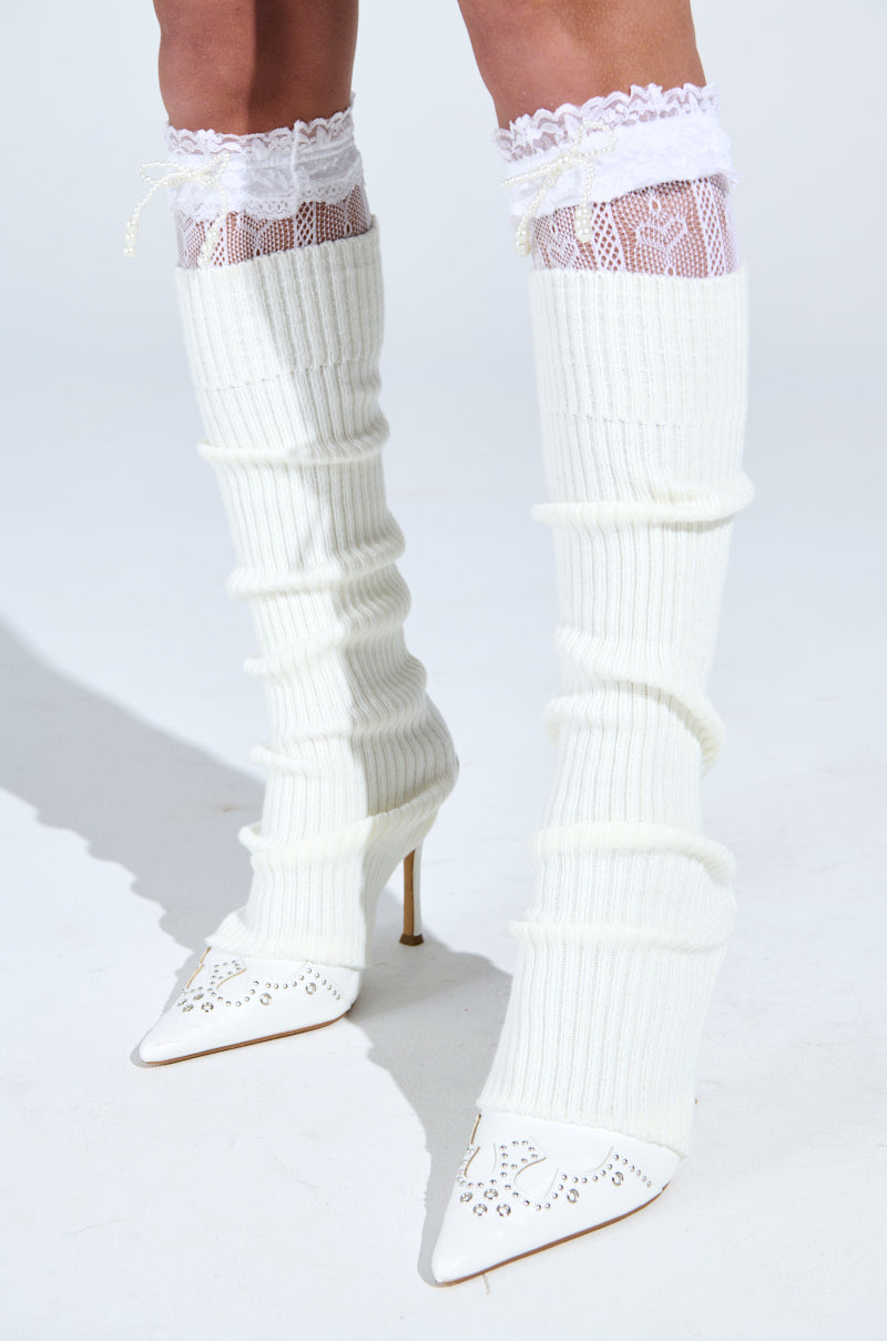 Wearing white knee-high COCO STOCKING LEG WARMERS over lace-patterned stockings captures a balletcore vibe, completed with elegant white stiletto heels with decorative cut-outs on a light background.