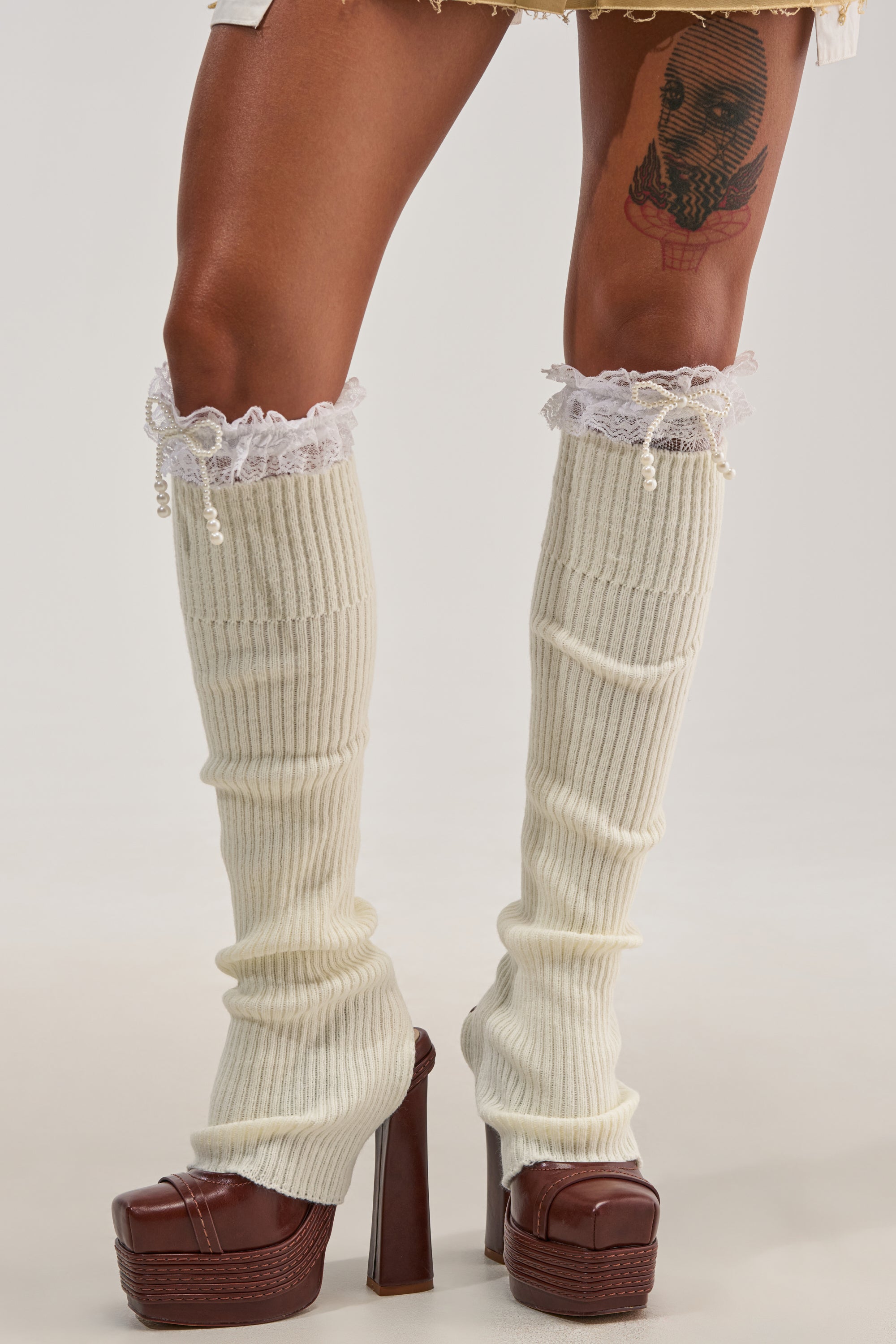 Wearing the COCO STOCKING LEG WARMERS in a cream hue, featuring delicate lace trim, this look pairs effortlessly with brown high-heeled platform shoes for winter-ready style. The ensemble is completed with a light-colored skirt and a thigh tattoo just visible above the leg warmers, infusing the outfit with unique personality and charm.