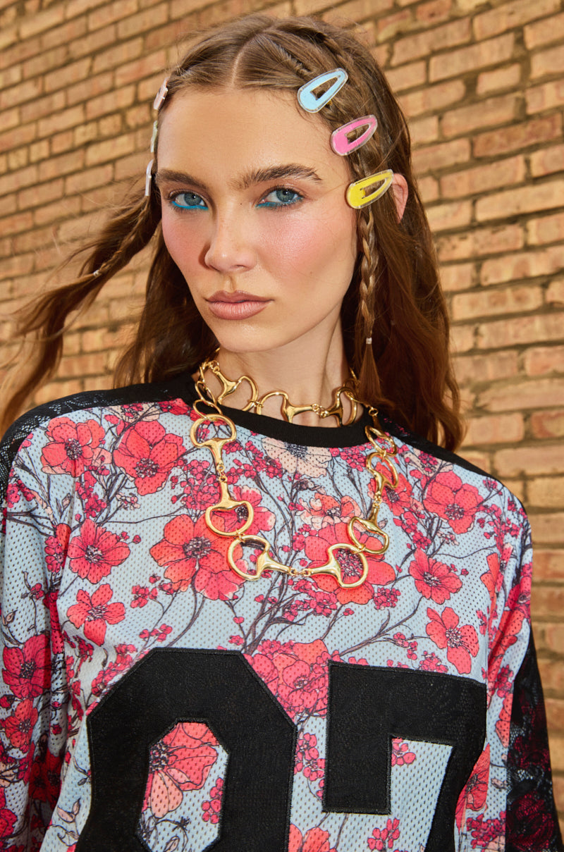 A woman with bold blue eyeliner, pink blush, and braided hair adorned with the IT'S A VISION HAIR CLIP SET wears a floral-patterned top and chunky gold necklace, standing against a brick wall.