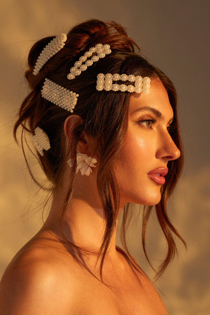 A brunette woman styled in an updo with the ROCKI HAIR CLIP SET, featuring pearl embellishments, looks aside. Her floral-shaped earrings and the warm, soft lighting beautifully highlight her features. 0