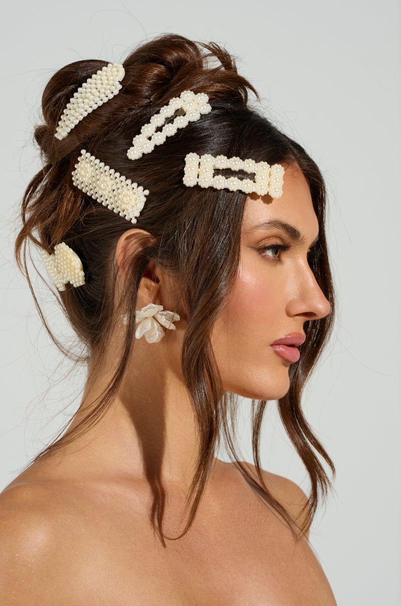 A woman with a stylish updo adorned with the ROCKI HAIR CLIP SET in pearls wears delicate translucent flower-shaped earrings against a plain, neutral background. 1