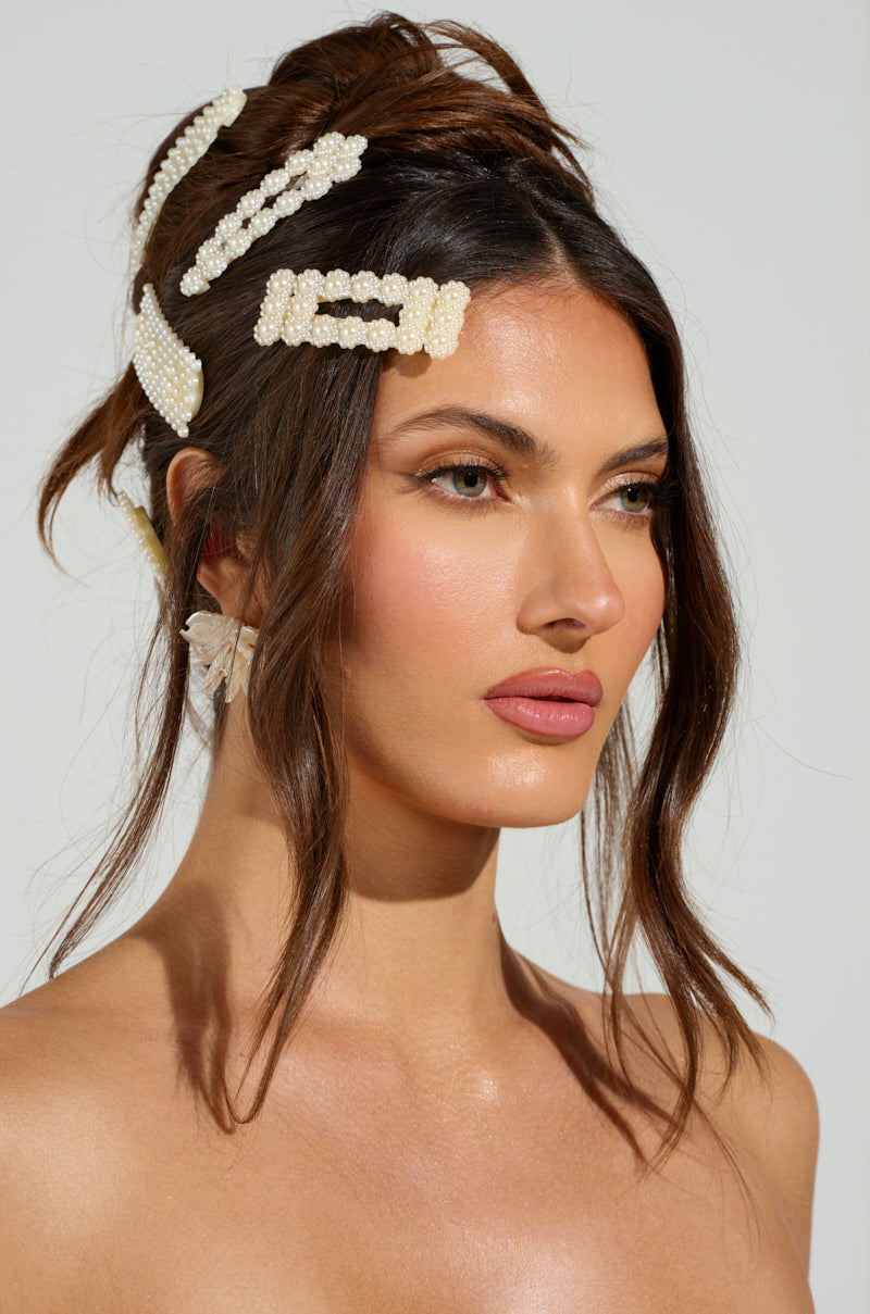 A woman with wavy brown hair, styled up with loose strands framing her face, wears subtle makeup. She's adorned with the ROCKI HAIR CLIP SET featuring pearl embellishments and a barrette-style clip, complemented by a large floral earring against a simple light-colored background. 2