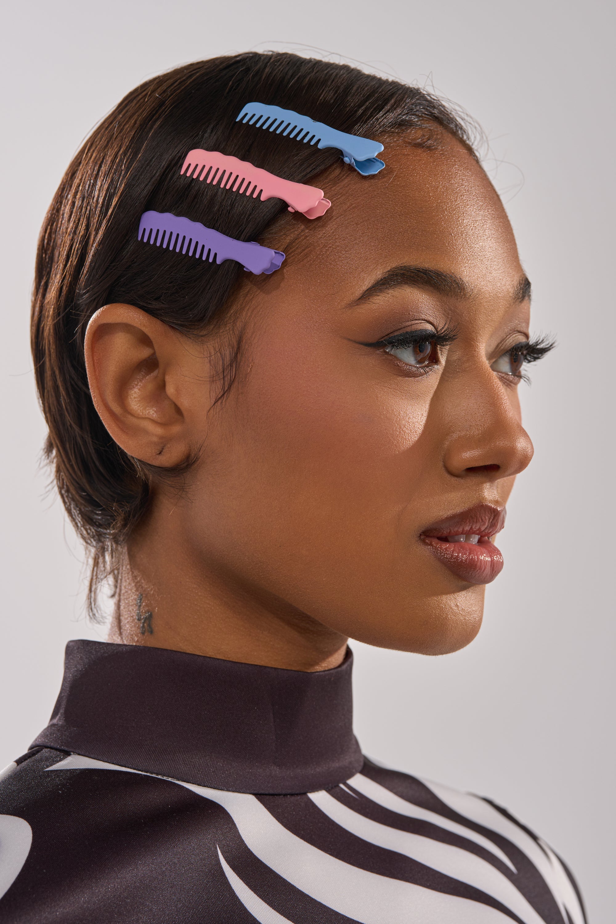 A woman with smooth, dark hair wears the COMB HERE HAIR CLIP SET—three pastel comb barrettes in purple, pink, and blue—on the side of her head. She sports winged eyeliner and a black-and-white patterned top. 0