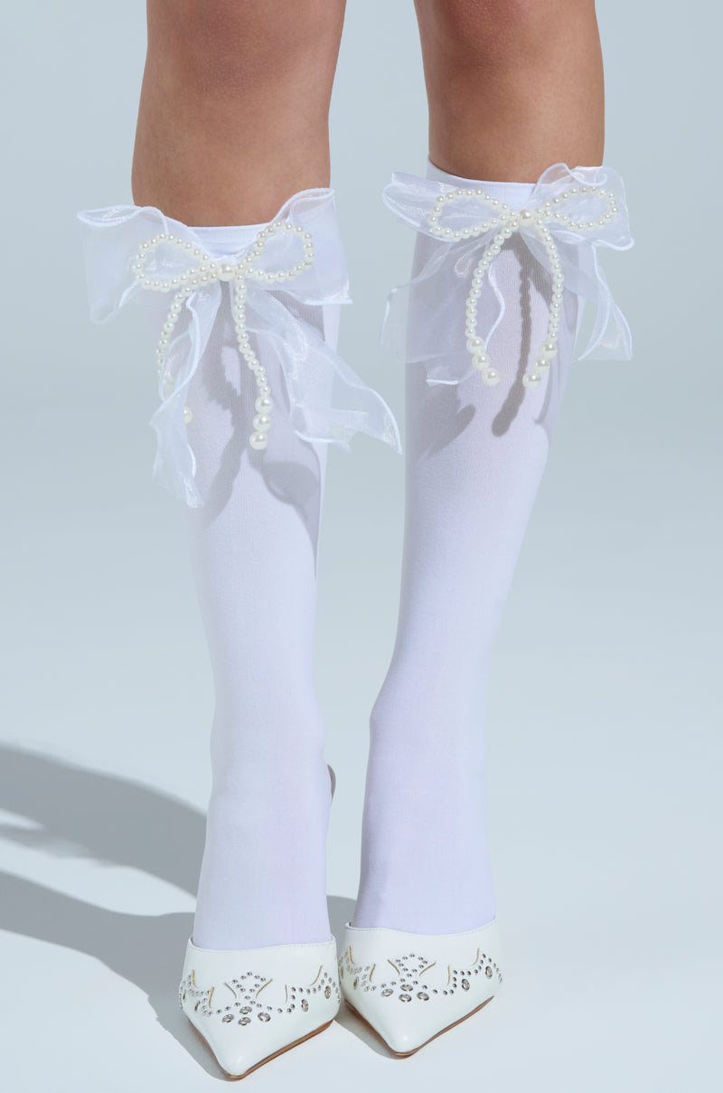 A chic woman showcases MOD BOW SOCKS, white knee-highs adorned with shimmery bows and pearls at the back, paired with white pointed heels featuring a perforated design, all against a serene light blue backdrop. 0