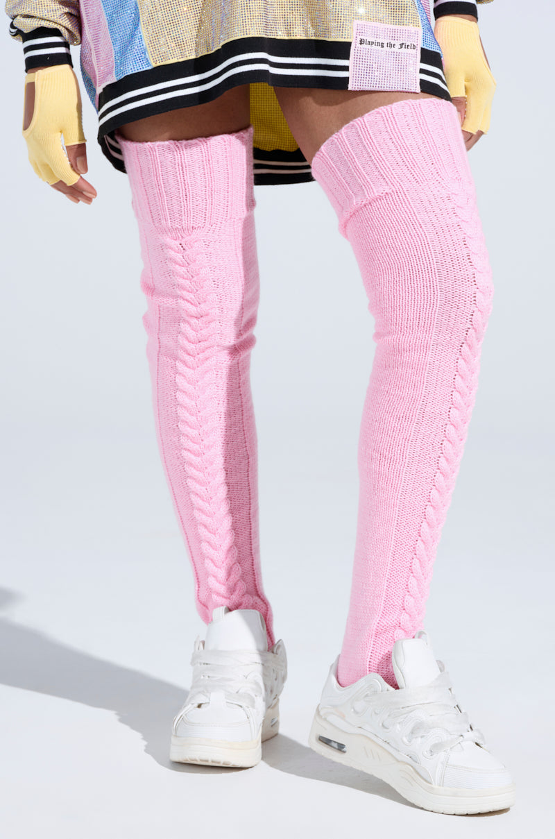 A person wearing STILL COZI THIGH HIGH SOCKS in cozy pink cable knit, white sneakers, a colorful skirt, and yellow fingerless gloves stands against a light background. 0
