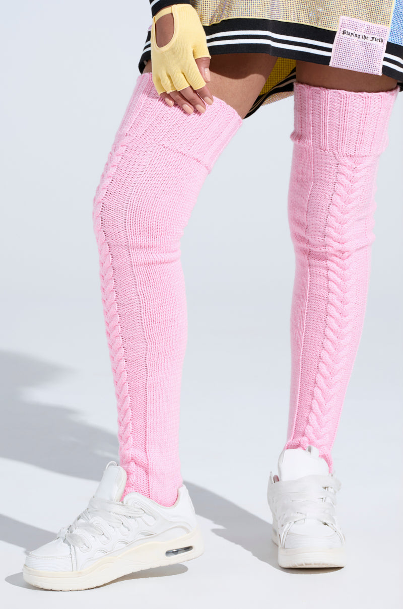 A person wearing STILL COZI THIGH HIGH SOCKS in cozy pink cable knit, paired with white sneakers, a beige skirt, and a yellow fingerless glove, stands against a white background.