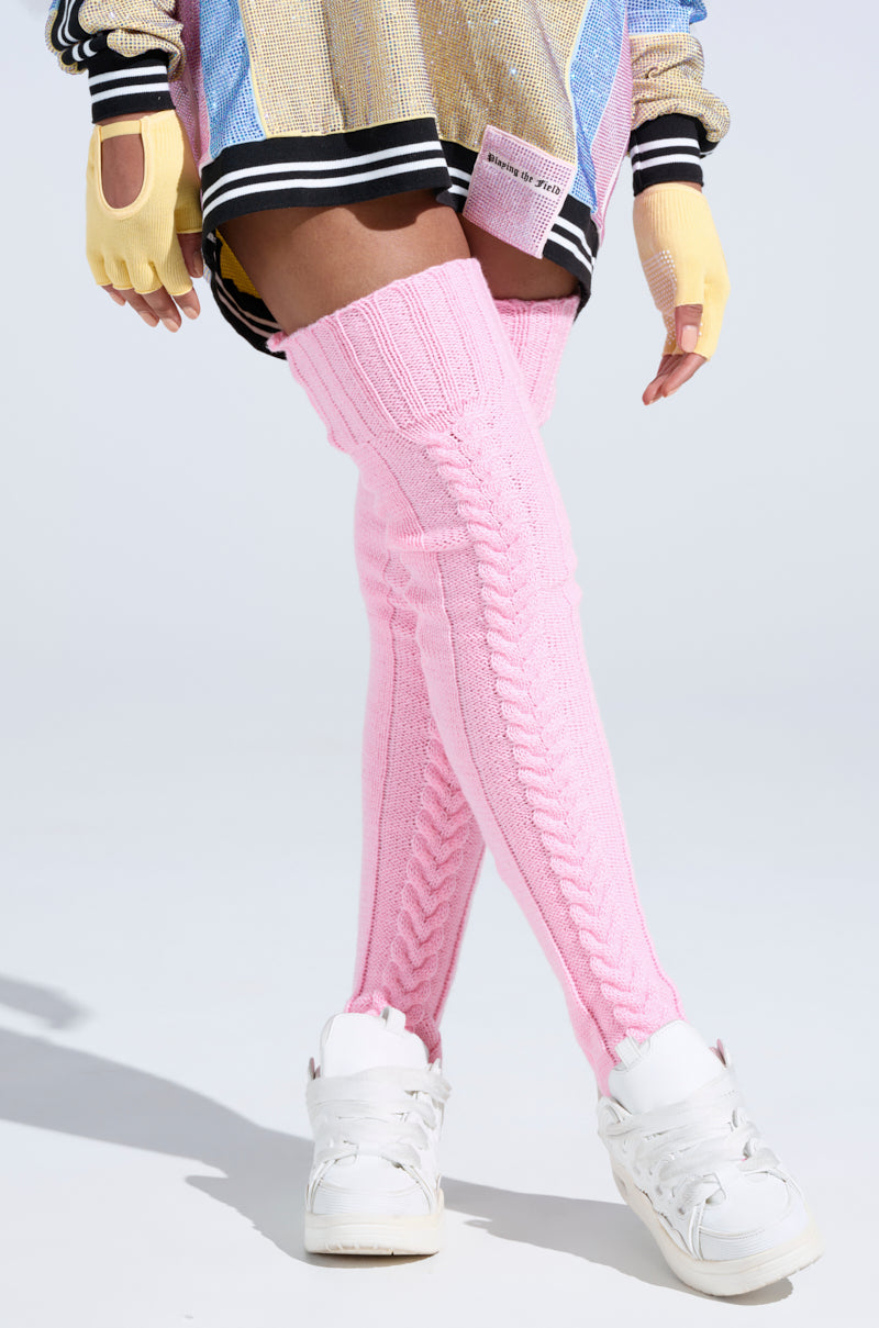 A person stands against a light background wearing colorful attire, yellow fingerless gloves, white sneakers, and STILL COZI THIGH HIGH SOCKS in cozy pink cable knit style.