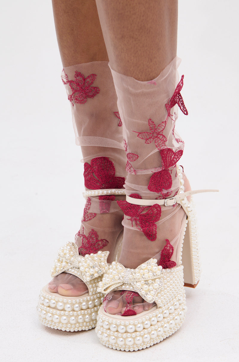 A close-up of a person wearing TAKE ME AWAY SOCKS—sheer pink with red floral embroidery—paired with platform heels adorned with pearls and bows against a white background. 0
