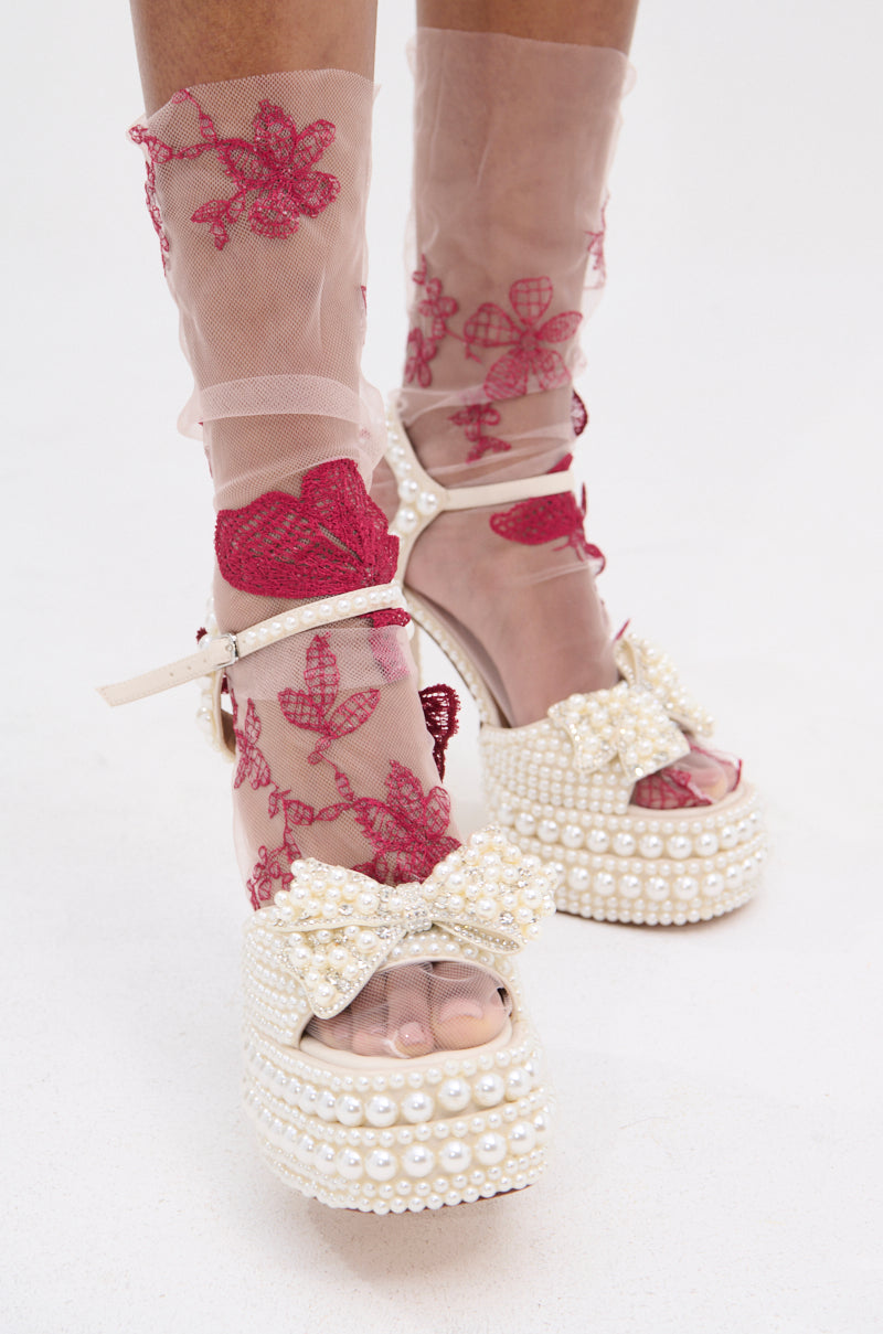 A person wears TAKE ME AWAY SOCKS—sheer pink socks with red floral embroidery—paired with white platform heels adorned with pearls and pearl bows, standing on a white surface. 2