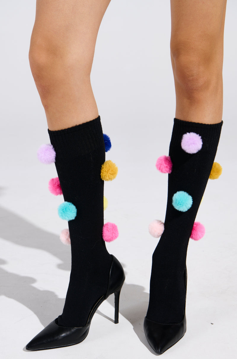 A pair of legs wearing black high-heeled shoes and knee-high HAVE A LIL FUN POM SOCKS, featuring colorful pom-poms in blue, pink, yellow, and purple. The playful accents add whimsy against a plain backdrop, highlighting the mid-calf silhouette with quirky charm. 0
