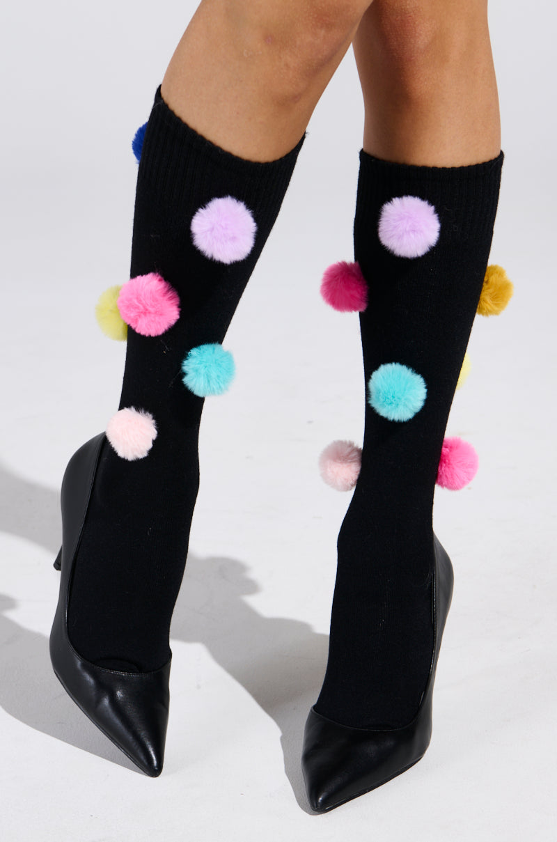 Black high-heeled shoes paired with HAVE A LIL FUN POM SOCKS featuring knee-length, black socks adorned with colorful pom-poms in pink, purple, blue, and yellow on a crisp white background, highlighting the playful footwear. 1