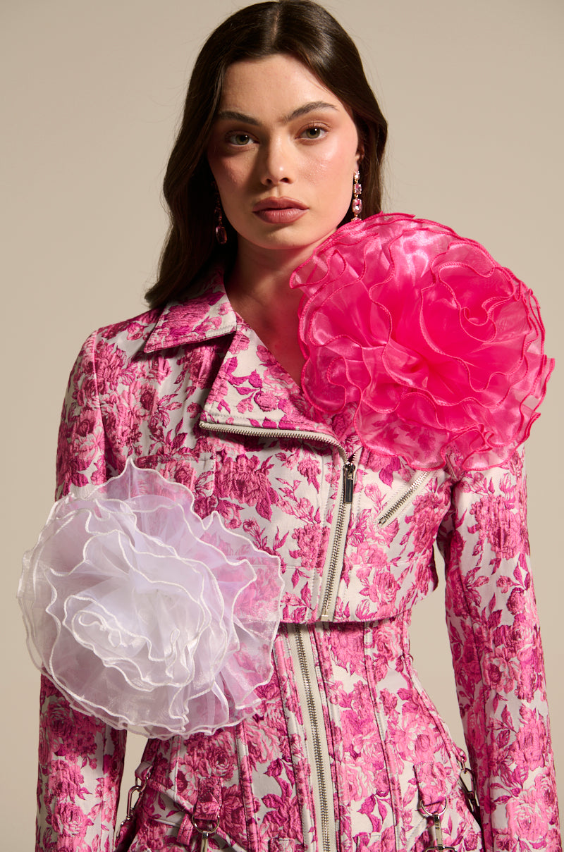A person in a floral-patterned pink jacket with oversized white and pink TIS THE SEASON BROOCH SET exudes avant-garde romance. Their long dark hair cascades against a neutral backdrop, enhancing the organza-like elegance.