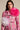 A person in a floral-patterned pink jacket with oversized white and pink TIS THE SEASON BROOCH SET exudes avant-garde romance. Their long dark hair cascades against a neutral backdrop, enhancing the organza-like elegance.