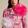 A person in a floral-patterned pink jacket with oversized white and pink TIS THE SEASON BROOCH SET exudes avant-garde romance. Their long dark hair cascades against a neutral backdrop, enhancing the organza-like elegance.