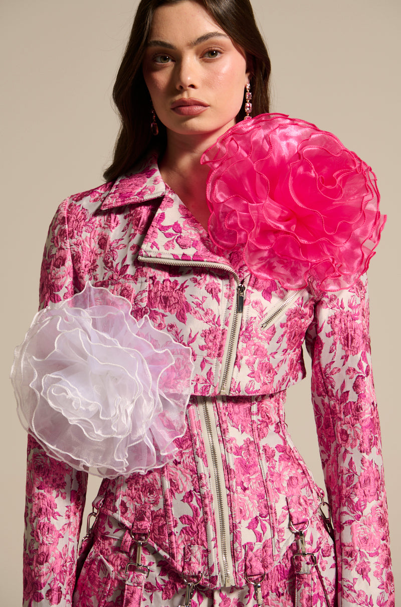 A person in a fitted, pink floral-patterned jacket adorned with TIS THE SEASON BROOCH SET featuring large ruffled ruffles and oversized pink and white fabric flowers. Zippers enhance the design, adding avant-garde romance against a neutral background.