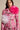 A person in a fitted, pink floral-patterned jacket adorned with TIS THE SEASON BROOCH SET featuring large ruffled ruffles and oversized pink and white fabric flowers. Zippers enhance the design, adding avant-garde romance against a neutral background.