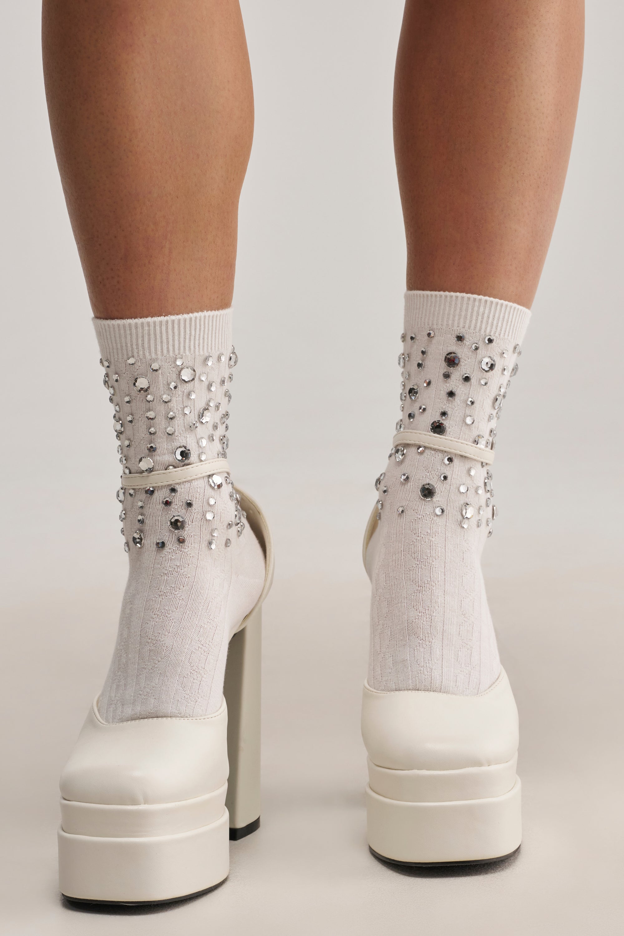 A close-up of legs wearing BLING BLING SOCKS—white embellished socks with silver studs—paired with white high-heeled platform shoes, shown from the back against a plain background. These socks are perfect festival accessories to make your outfit shine.