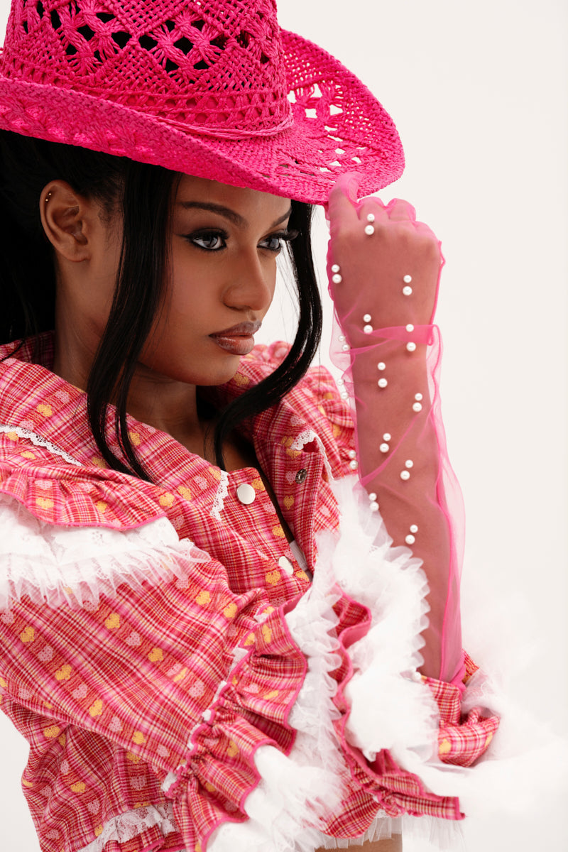 A woman poses confidently in a pink plaid outfit with ruffled white trim, wearing a bright pink cowboy hat and ALL THE VIBES PEARL GLOVES IN PINK—sheer tulle gloves embellished with pearls—as she looks to the side holding her hat’s brim. 1