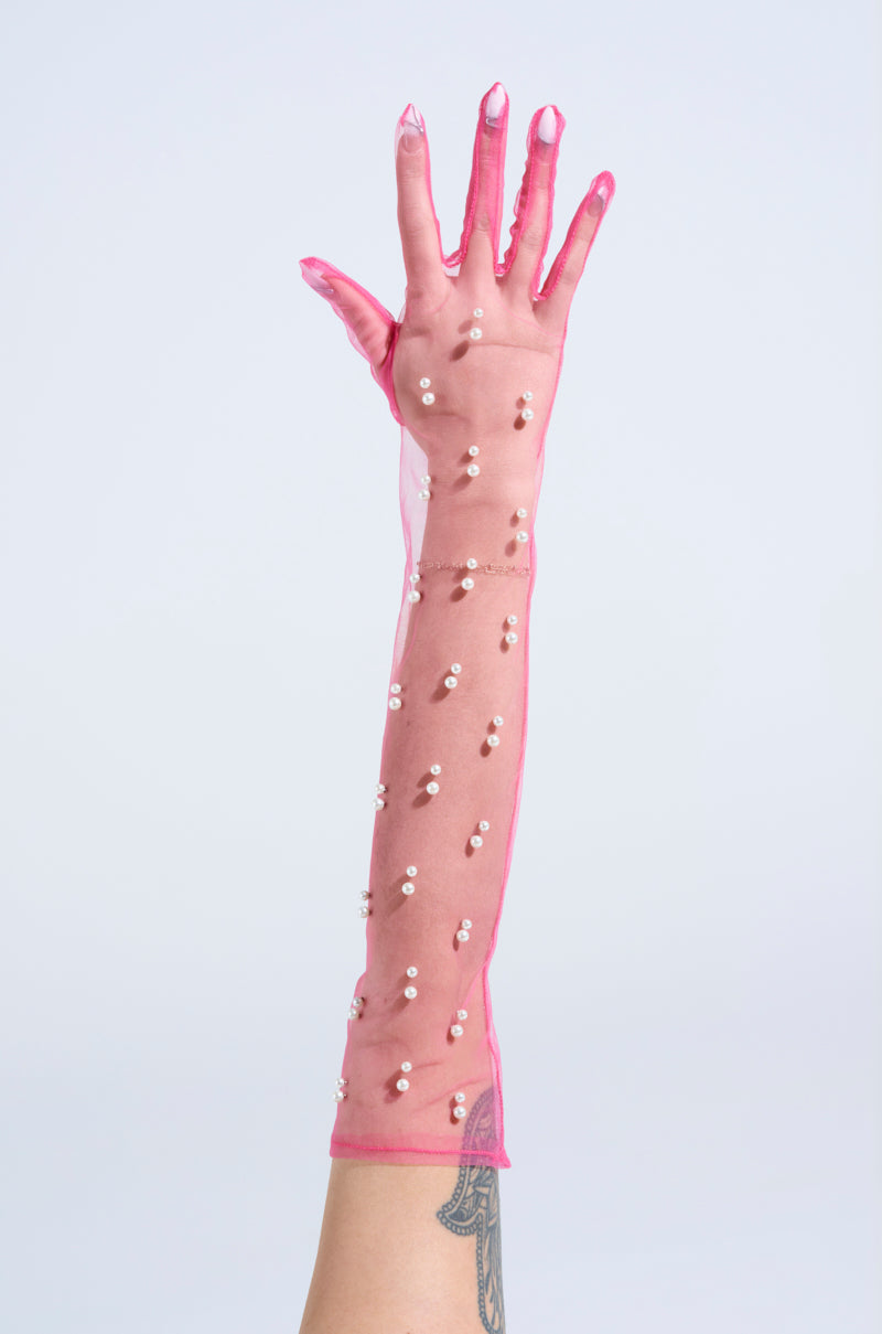 A hand wearing the ALL THE VIBES PEARL GLOVES IN PINK, made of sheer tulle and adorned with pink pearls, is raised against a light background, revealing the wearer’s tattooed forearm through the embellished glove. 2