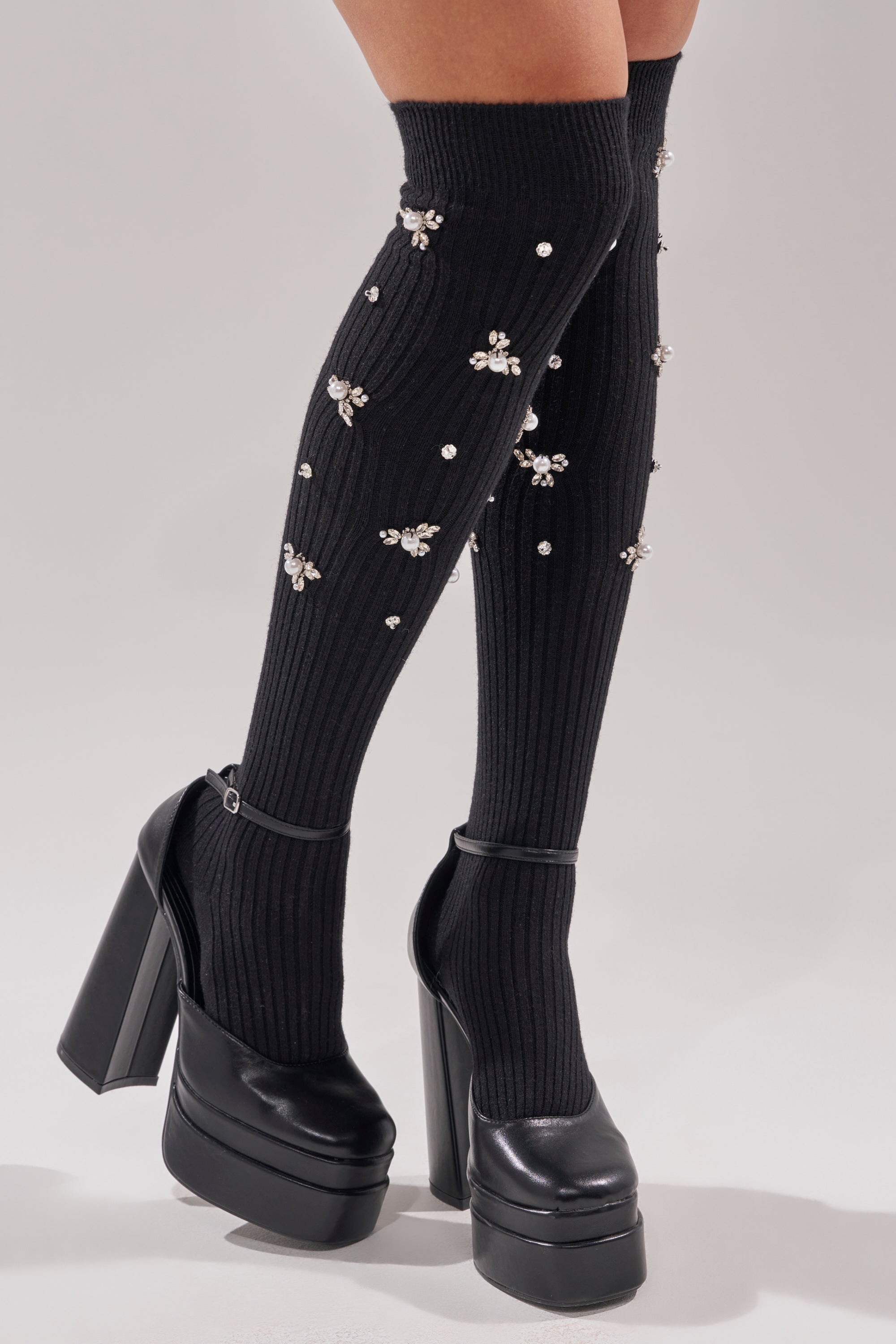 A person wears COCO EMBELLISHED SOCKS—ribbed, black, over-the-knee socks adorned with rhinestone flowers—paired with black high-heeled platform Mary Jane shoes against a plain light background. 0