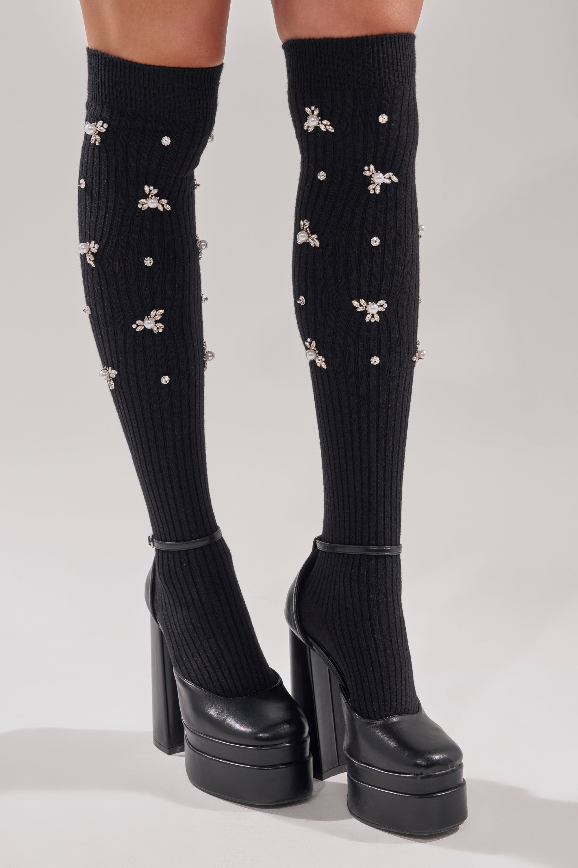A person wears COCO EMBELLISHED SOCKS, featuring black fabric decorated with small rhinestone flowers, styled with black high platform heels against a plain background. 1
