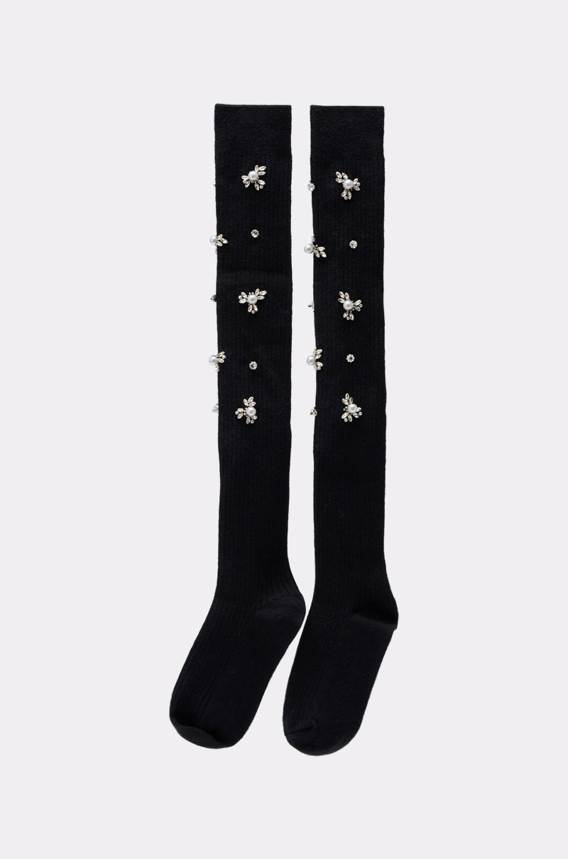The COCO EMBELLISHED SOCKS are black over-the-knee socks adorned with sparkling rhinestone floral designs running vertically along the front, ideal for adding a touch of glamour to your look. 2