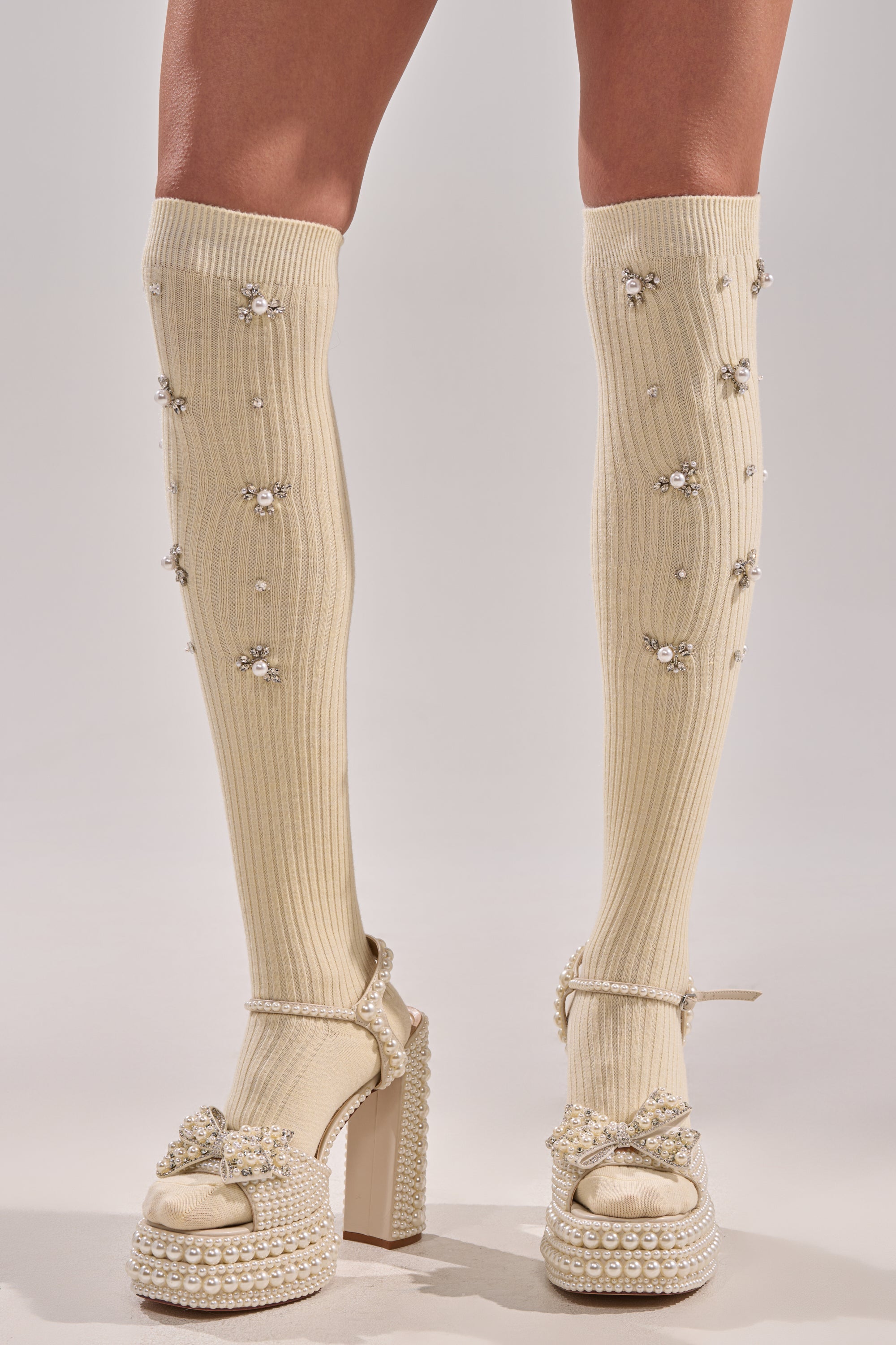 A pair of legs styled in COCO EMBELLISHED SOCKS IN CREAM, featuring knee-high cream socks with small gems, paired with chunky high-heeled sandals adorned with pearls and jeweled details for a chic embellished look. 0