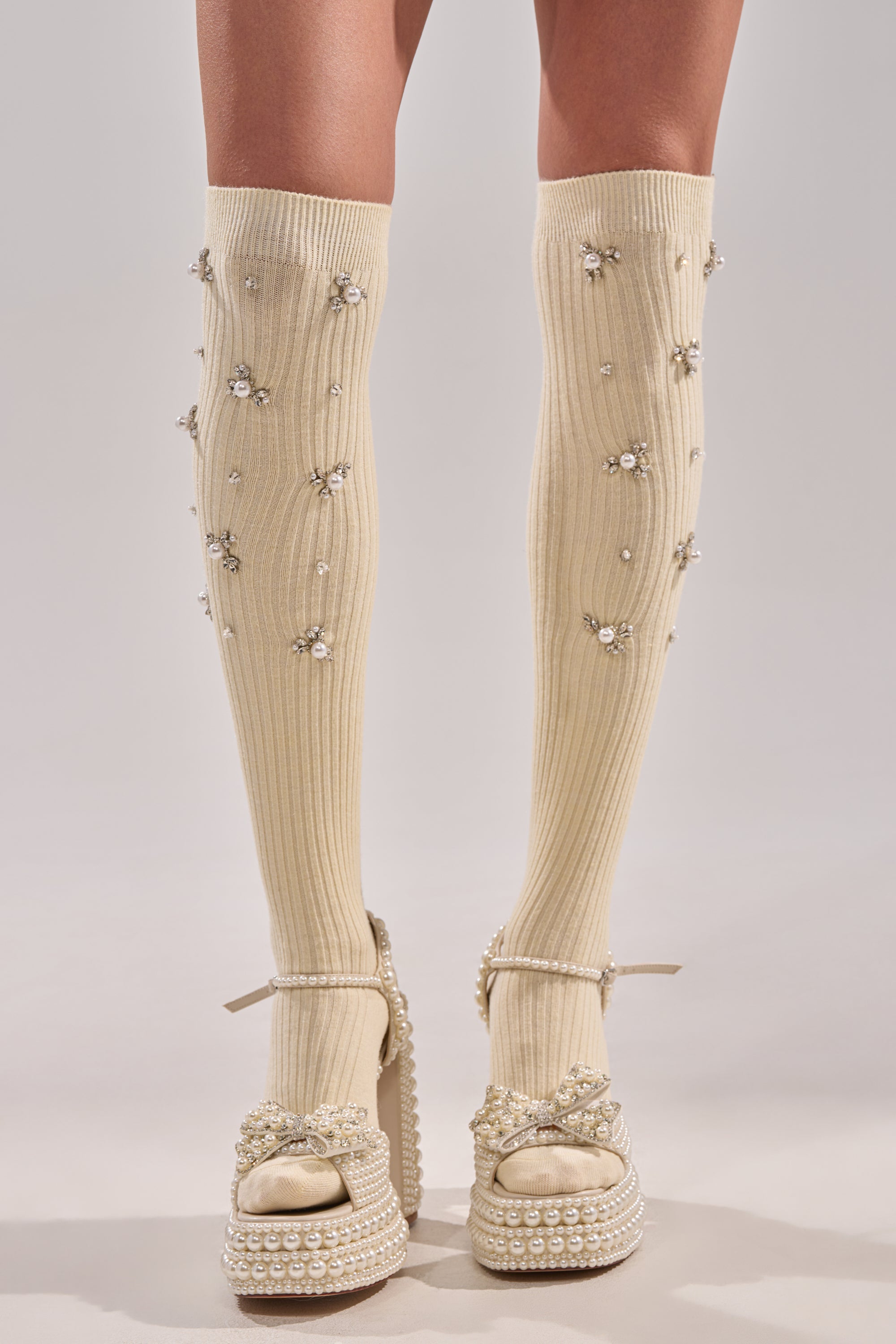 A person wears COCO EMBELLISHED SOCKS IN CREAM—knee-high and adorned with decorative pearls and rhinestones—paired with high platform pearl-embellished sandals. Only the legs and feet are visible, emphasizing these elegant socks. 3