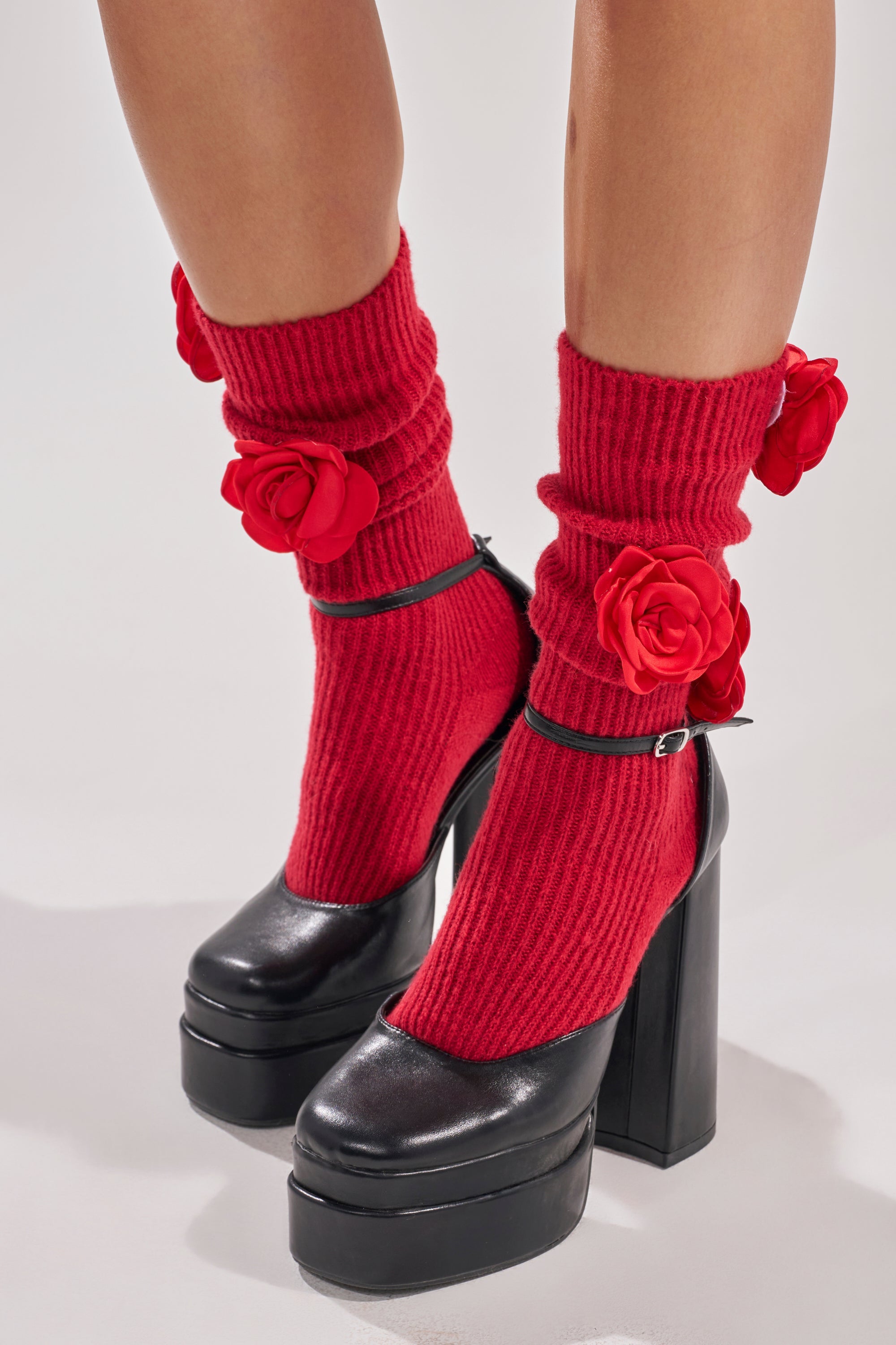 Someone wears ROSIE SOCKS—bright red, mid-calf socks with ruffled edges and rosette appliques—paired with chunky black high-heeled Mary Jane shoes. The background is plain and light-colored. 0