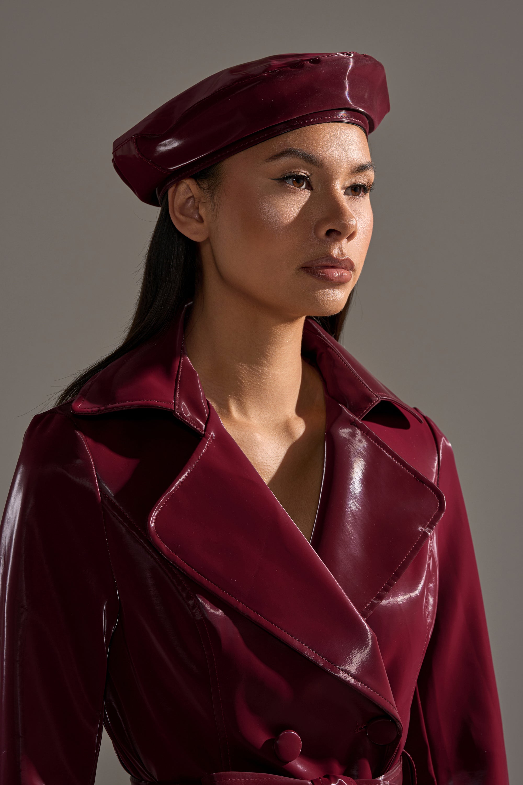 A woman in a glossy burgundy trench coat and the LIQUID VELVET BERET IN BURGUNDY stands against a neutral background, gazing slightly aside with a serious expression. 2