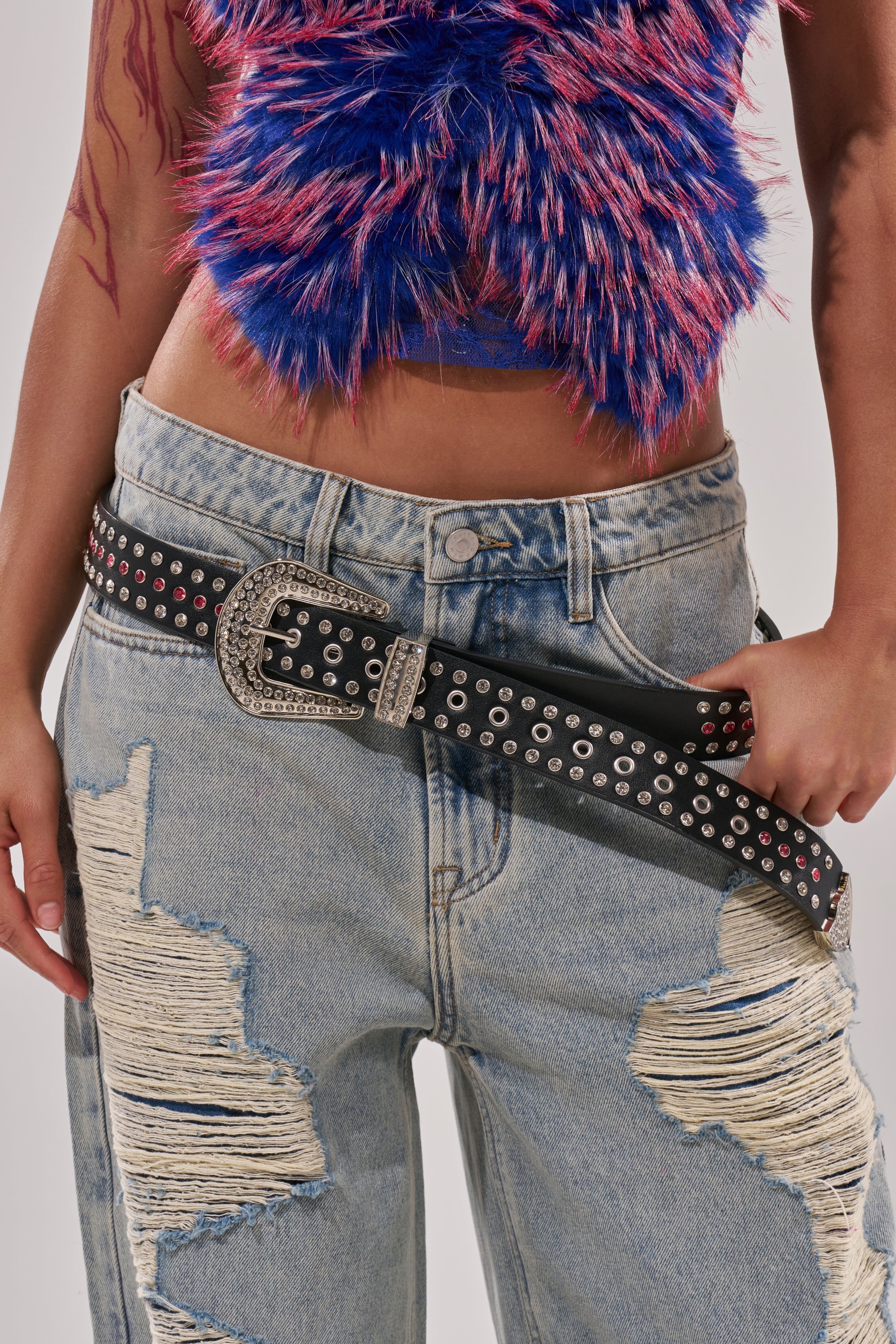 Wearing distressed light blue jeans and a fuzzy blue and pink crop top, the person shows off the I AM THAT BELT—a crystal-studded belt with a large western buckle—with their hand resting near it. 9
