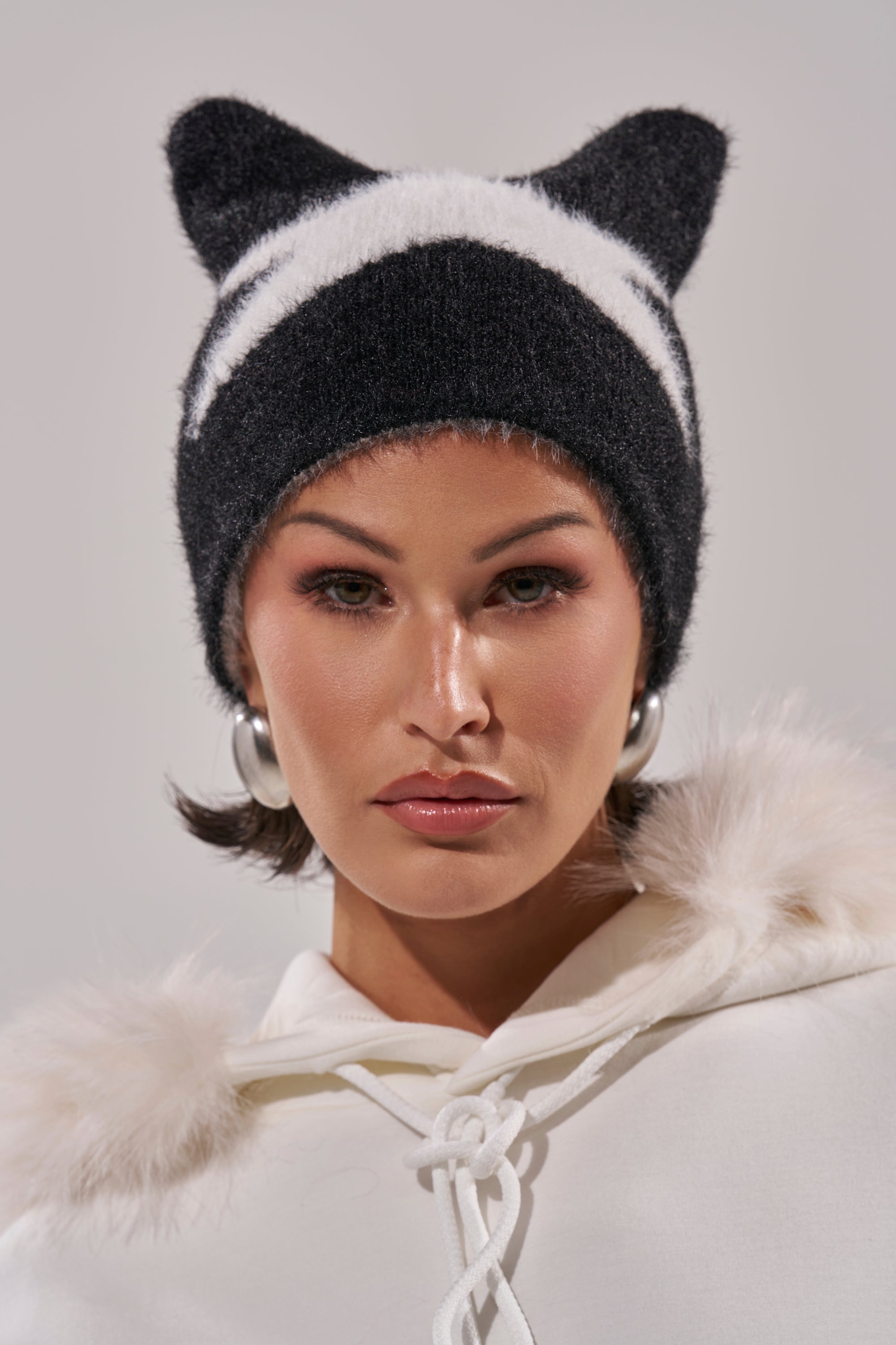 A woman wears the IT GURL BEANIE—a black and white knit hat with cat-like ears—paired with a faux fur-trimmed white hoodie and silver hoop earrings, looking directly at the camera against a plain background. 4