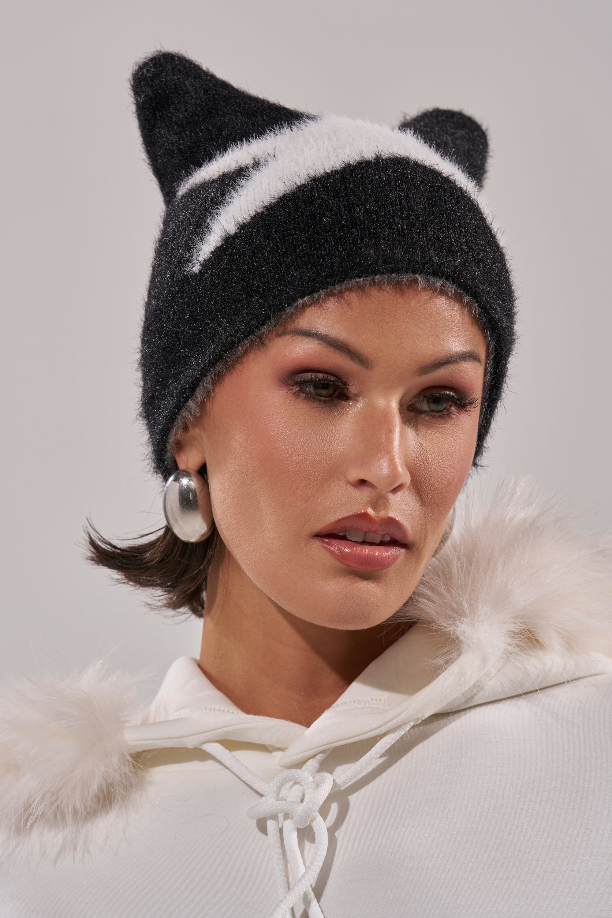 A woman wears the IT GURL BEANIE, a black and white knit hat with cat-ear shapes, paired with a white faux fur-trim hoodie. She has hoop earrings, natural makeup, and gazes slightly downward. 6