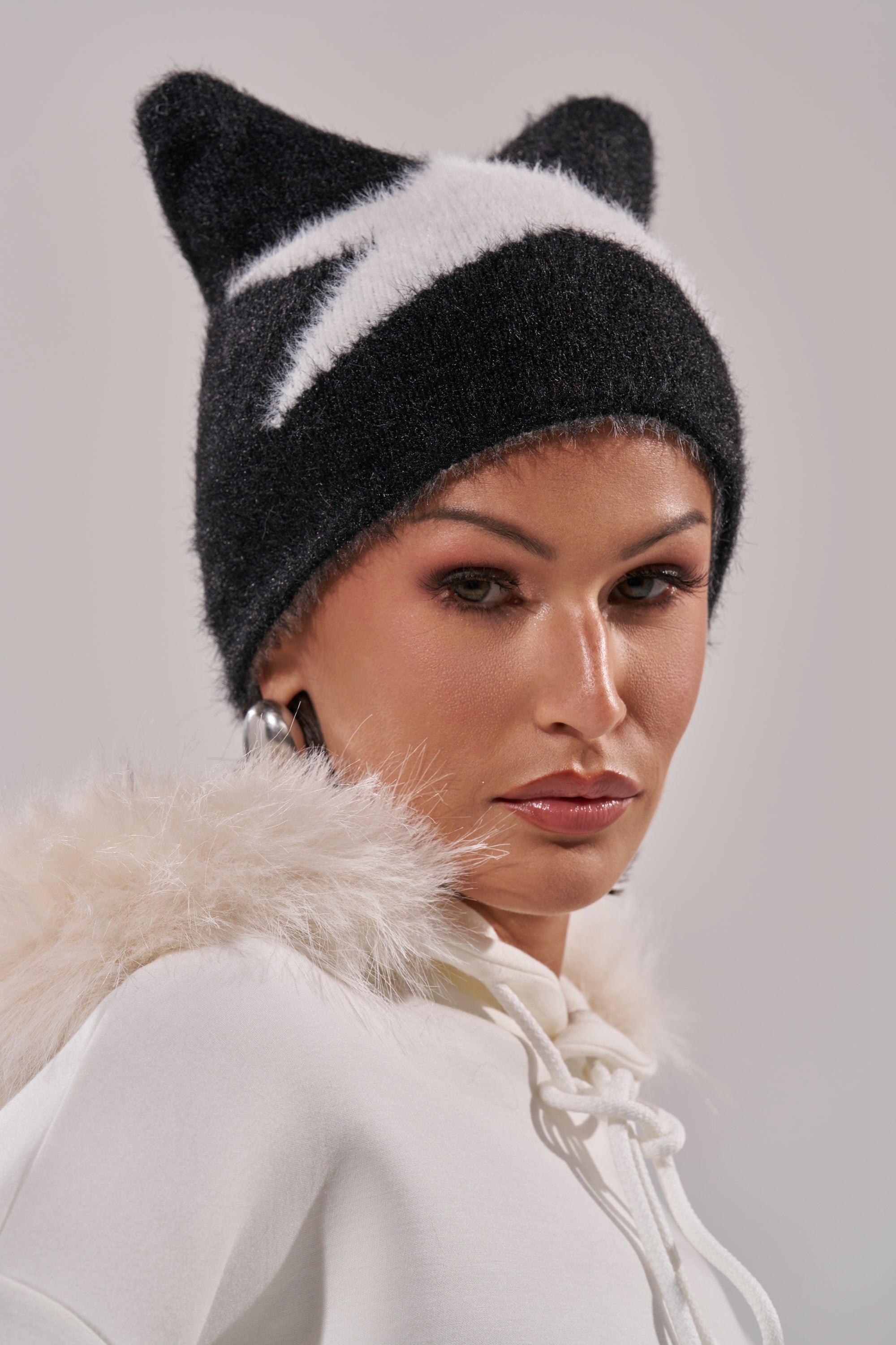 Wearing the IT GURL BEANIE with cat-like ears and white star accents, paired with a white hoodie with a fluffy hood trim, the person glances over their shoulder with a neutral expression. 7